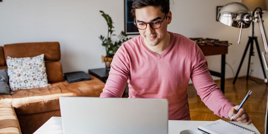 Junger Mann arbeitet im Homeoffice am Laptop, macht Notizen in ein Notizbuch, moderner Arbeitsplatz mit Sofa und Pflanzen im Hintergrund.