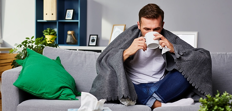 VRG HR Blog_Krank im Urlaub Erkälteter Mann auf dem Sofa mit Tasse und umgeben von benutzten Taschentüchern